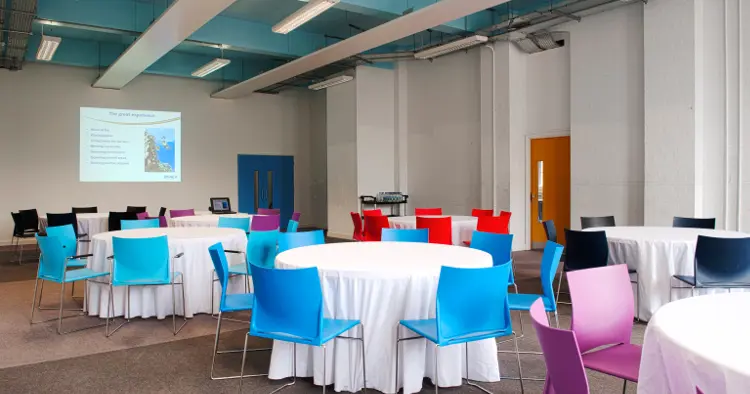 East side of blue hall with round tables with white table cloths and colourful chairs. Projection on to wall at end.