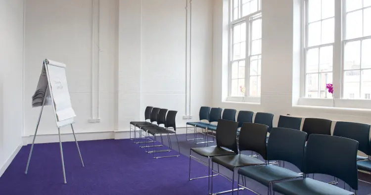 Meeting room with white walls and purple carpet and two large Georgian style windows. 16 chairs are set in two rows and a flip chart at front.