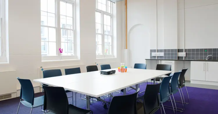Meeting room with white walls and purple carpet and two large Georgian style windows. 12 chairs are set round a white board room table. There is a sink unit in background.