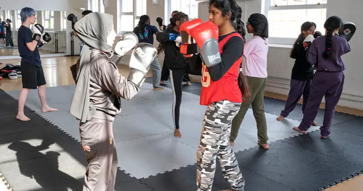 In the foreground two young women wearing boxing gloves are sparring, other young people in the background