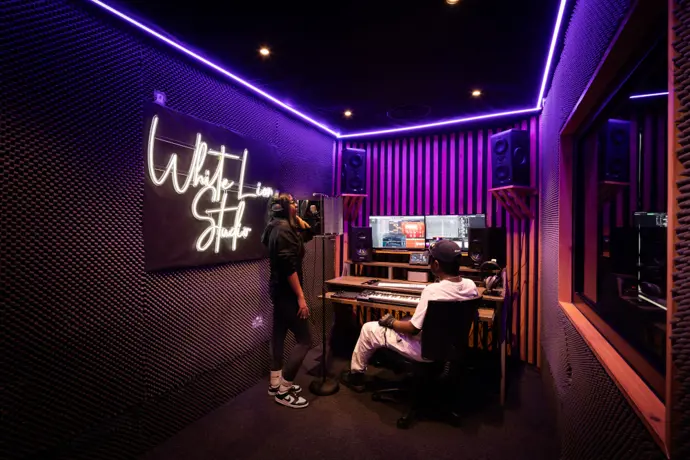 Recording studio with desk, large speakers and computer screens showing music software. Man stitting at desk and woman standing with headphones on
